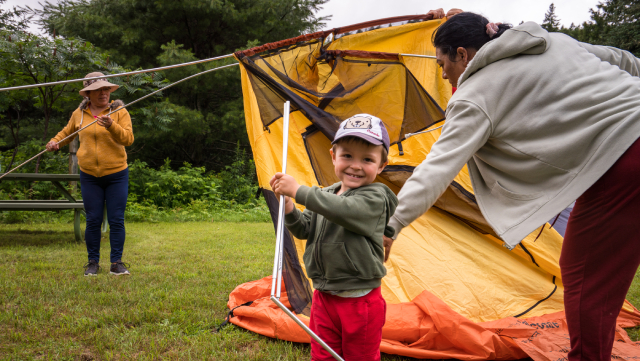 Two adults and a young child learn to set up a tent on a cloudy day on a grassy area.