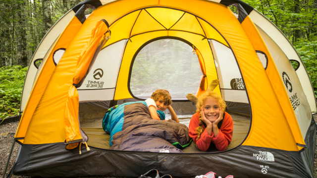 Two children pose for a photo, lying down inside of a yellow tent in a forest. 