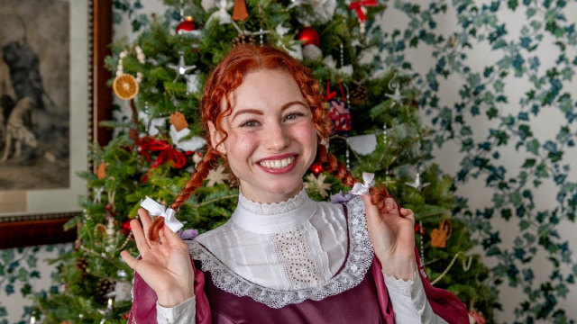 Une guide de Parcs Canada vêtue d'une tenue du 18e siècle représentant Anne de la maison aux pignons verts tient ses tresses et sourit à la caméra devant un sapin de Noël.
