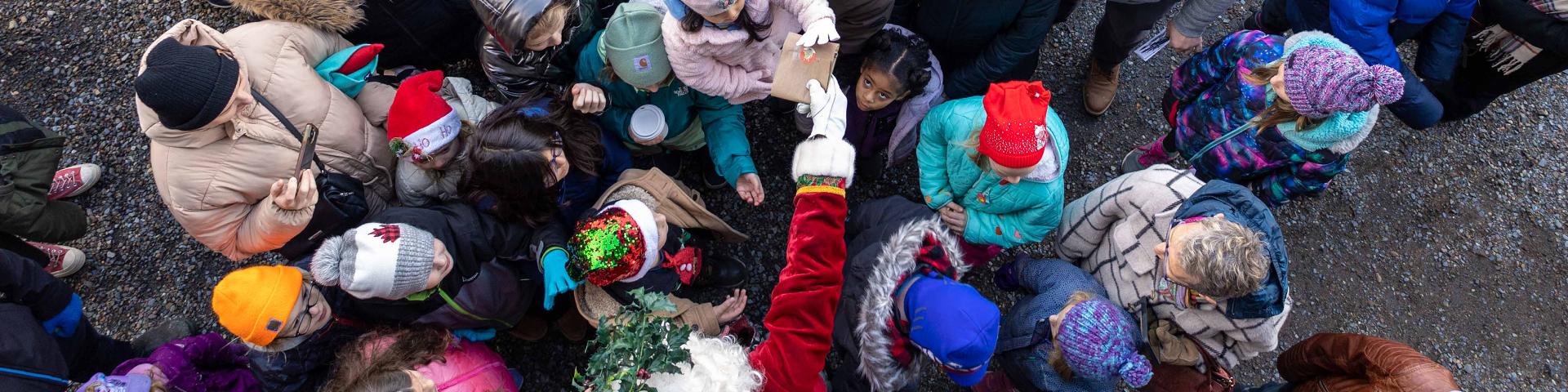 An aerial shot of santa handing out presents to a large group of people in winter outdoor coats. 