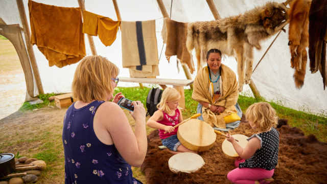A woman takes a photo of her two children who sit in a drum circle with an Indigenous person leading an activity in a tipi with furs.