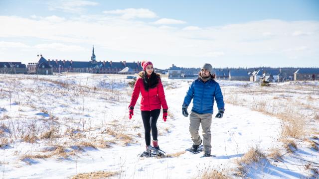 Deux visiteurs font de la raquette sur un sentier enneigé avec la forteresse de Louisbourg en arrière-plan.