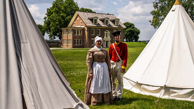 Two staff member dressed in period costume stand in between historic canvas tents.