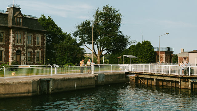 A family of three stands along the side of historic canal with a red brick building in the background. 