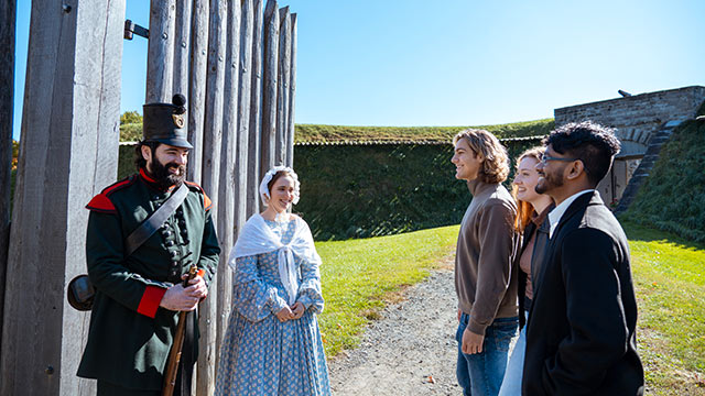 Three visitors stand at a wooden gate with two staff members in period costume. 