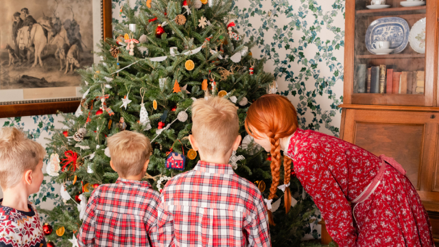 A costumed interpreter dressed as Anne of Green Gables looks at a Christmas tree with three boys in festive clothing.