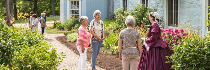 Des visiteurs discutent avec un guide en costume d’époque devant les jardins du manoir Papineau par une journée ensoleillée.