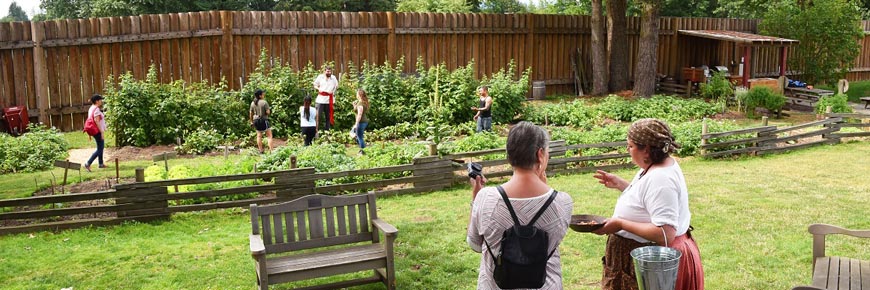 A visitor speaking with a gardener in historic costume in front of Fort Langley’s heritage garden as another guide talks with children in the garden.