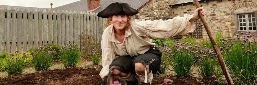 A guide in a historic costume sits in a heritage garden with an 18th century gardening tool at De la Plagne house. 