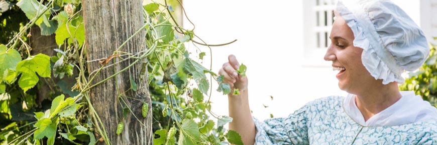 A Parks Canada guide in period costume tends to her garden with a smile on her face on a sunny day.  