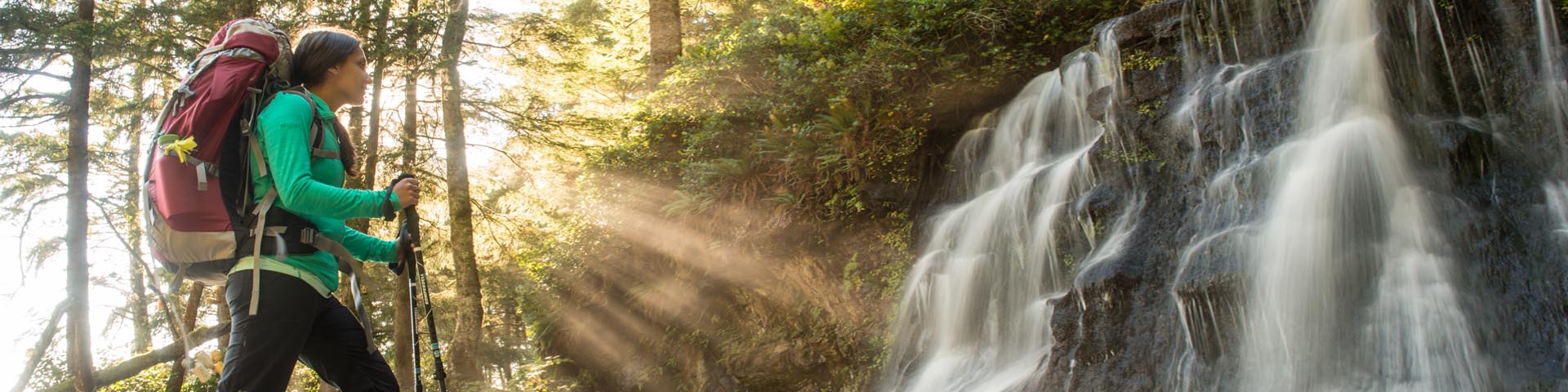  A female hiker marvels at the picturesque waterfall at Bonilla Creek, on the West Coast Trail, as the sun’s rays shine through the trees. Pacific Rim National Park Reserve.