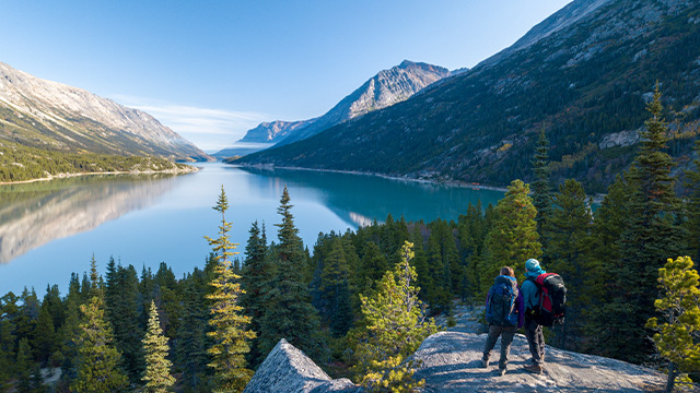 Two hikers with backpacks on the Chilkoot Trail, with a lake and mountains in the background.