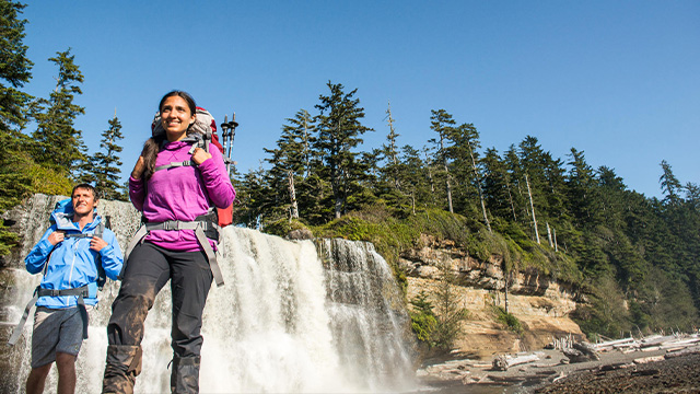 Two hikers walk along the beach beside the spectacular Tsusiat Falls in Pacific Rim.