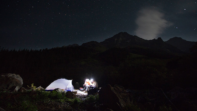Two campers beside a tent illuminated from within under a starry sky, with mountains in the background at Yoho.