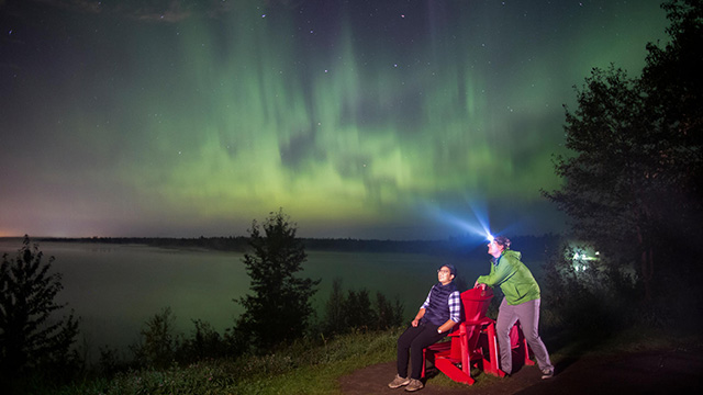 	
Un couple près de chaises rouges observe les aurores boréales à l’aire récréative du lac Astotin, dans le parc national Elk Island.