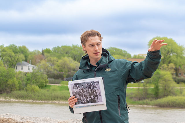 Un employé de Parcs Canada tient une photo et pointe à gauche au lieu historique national de La Fourche.