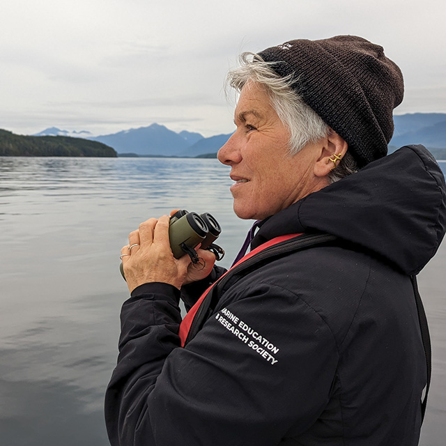 Carlo Acuna is wearing a green hoodie and black sunglasses. He is standing in front of the ocean