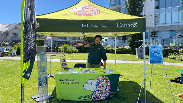 A Parks Canada tent on a grassy field. A Southern Resident Killer Whale team member is standing behind a green table in the Parks Canada uniform waving at the camera. Beside the table there is a brochure rack, a killer whale flag, and a map of the gulf islands.