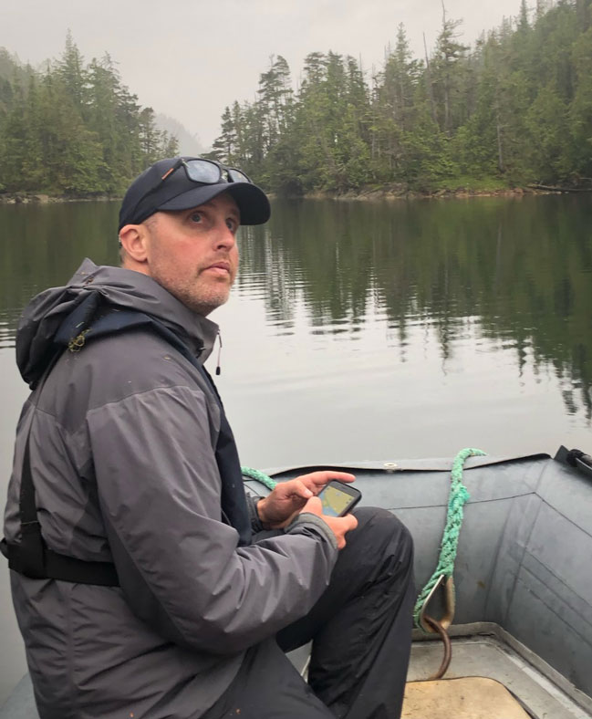 Gary in a boat on calm water. He is wearing rain gear and holding a phone
