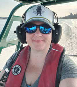 Ashley on a boat smiling at the camera. She has a red lifejacket on, hearing protection, sunglasses, and a hat with a bear on it. 