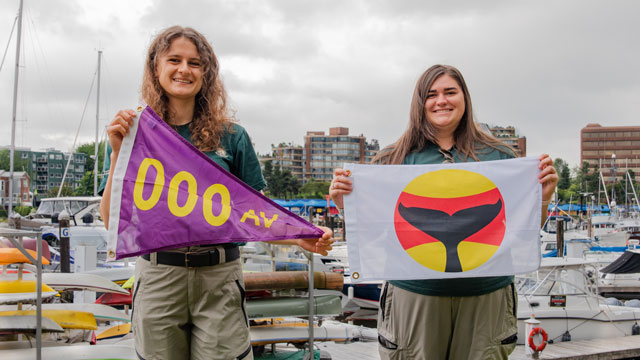 Deux membres du personnel de Parcs Canada brandissent des pavillons. Le pavillon de gauche, violet et triangulaire, signale les navires autorisés. Le pavillon de droite est carré et blanc avec un cercle rouge et jaune et une queue de baleine noire à l’intérieur du cercle. Il s’agit du pavillon d’avertissement de la présence de baleines. 