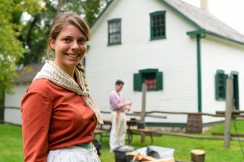A female interpreter in period costume in the foreground with Riel House and a male interpreter in the background.