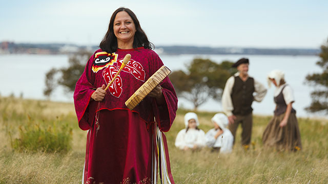 A woman in Mi’kmaw traditional dress stands in a grassy field holding a hand drum and drumstick, with body of water in the background and a family in historical clothing behind her.