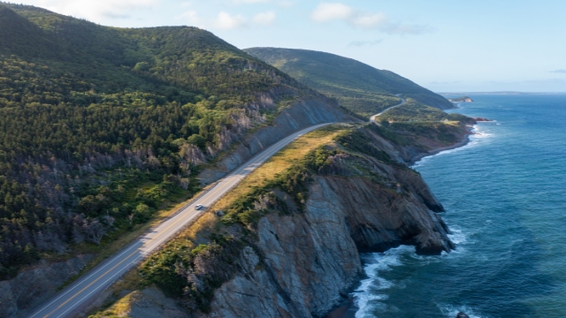 An aerial view of the Cabot Trail nestled between lush green mountains and the bright blue ocean and sky. A vehicle drives south on the right side of the road.