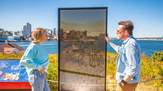 Two visitors look at a transparent glass panel depicting an historical moment at this location.