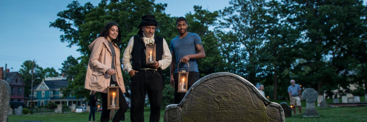 Un jeune couple et un guide costumé au crépuscule avec des lanternes sont devant une pierre tombale dans le cimetière de garnison.