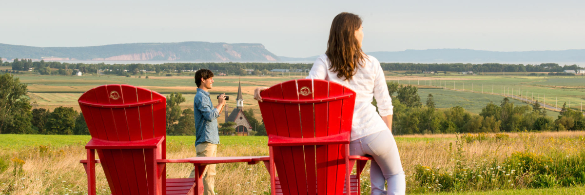 A couple next to red chairs in front of the Grand-Pré landscape