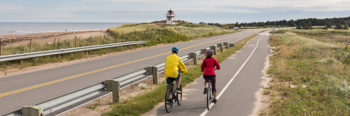 Deux cyclistes sur la piste le long de la route du Golfe avec un phare en arrière-plan au parc national de l’Île-du-Prince-Édouard.