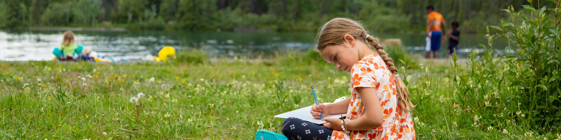 Une jeune fille assise dans l’herbe écrit dans un cahier, le lac Kathleen, des visiteurs et des montagnes en arrière-plan, au parc national et réserve de parc national Kluane.