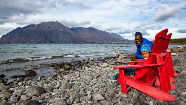 A man and a child sit on Parks Canada’s red chairs by a lake, in Kluane National Park and Reserve.