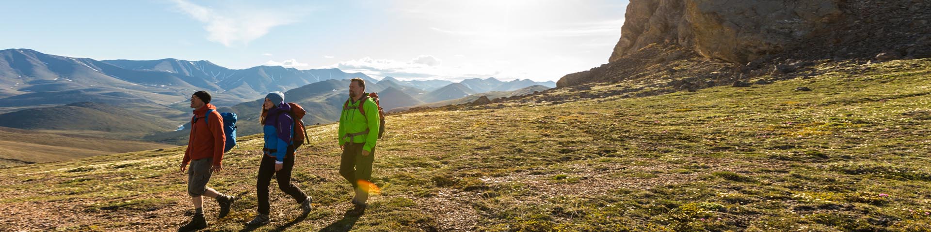 Trois visiteurs font de la randonnée dans la toundra avec le soleil et un rocher en arrière-plan au parc national Ivvavik.