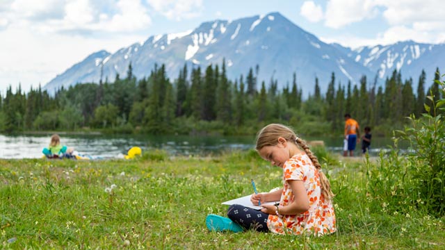 A young girl sitting in the grass writes in a notebook, Kathleen Lake, visitors and mountains in the background, at Kluane National Park and Reserve.