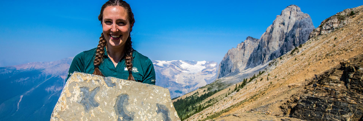 Une guide de Parcs Canada tient un fossile à la carrière Walcott, avec une vue du lac Emerald, du glacier Emerald et du sommet du mont Wapta.