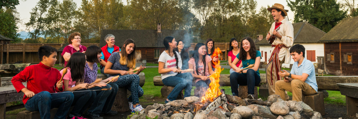 Visitors and a costumed guide sitting by a fire inside the palisades of Fort Langley.
