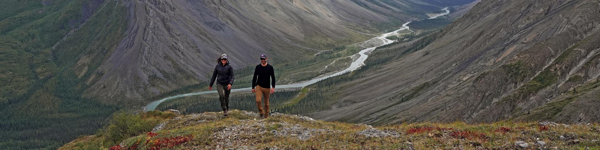 Une femme et un homme rient et marchent vers l’appareil photo, avec une vue d’une vallée fluviale et des montagnes environnantes en arrière-plan.