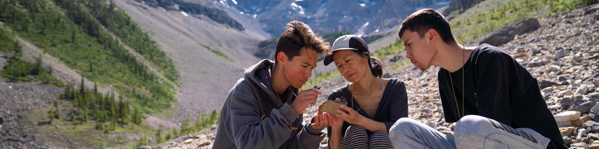 A woman and two young boys grouped together, crouch on rocky ground surrounded by mountains. They examine a fossil using a magnifying glass. 