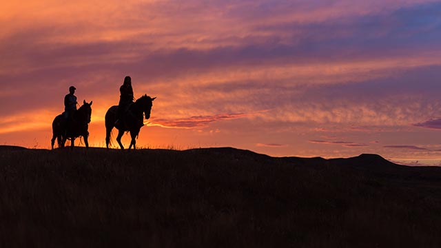 Two riders on horseback against the light of a bright pink and orange sunset at Grasslands National Park.