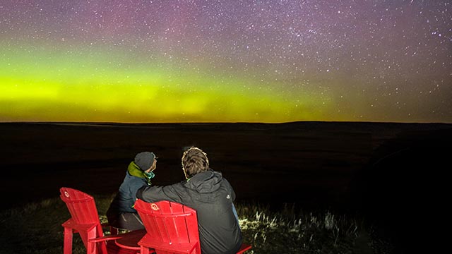 Deux visiteurs, confortablement assis sur des chaises rouges admirent de magnifiques aurores boréales vertes et mauves et la Voie lactée au parc national des Prairies.