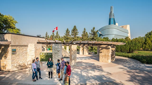 Un interprète de Parcs Canada guide les visiteurs pendant une visite historique près du Cercle d’orientation. On aperçoit le Musée canadien pour les droits de la personne en arrière-plan.