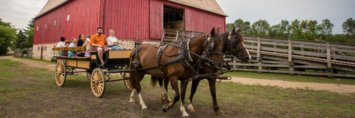 A team of horses pulls a wagon full of visitors past a big red barn.