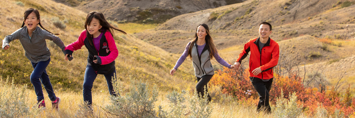 Two children running up a hill, with their parents following, with the landscape of the West Block of Grasslands National Park in the background.