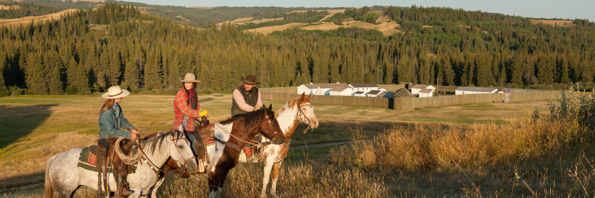 Three horseback riders discuss with Fort Walsh in the background. 