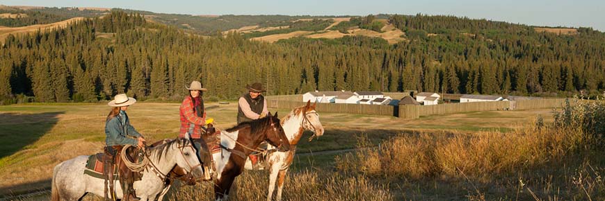 Three horseback riders discuss with Fort Walsh in the background. 