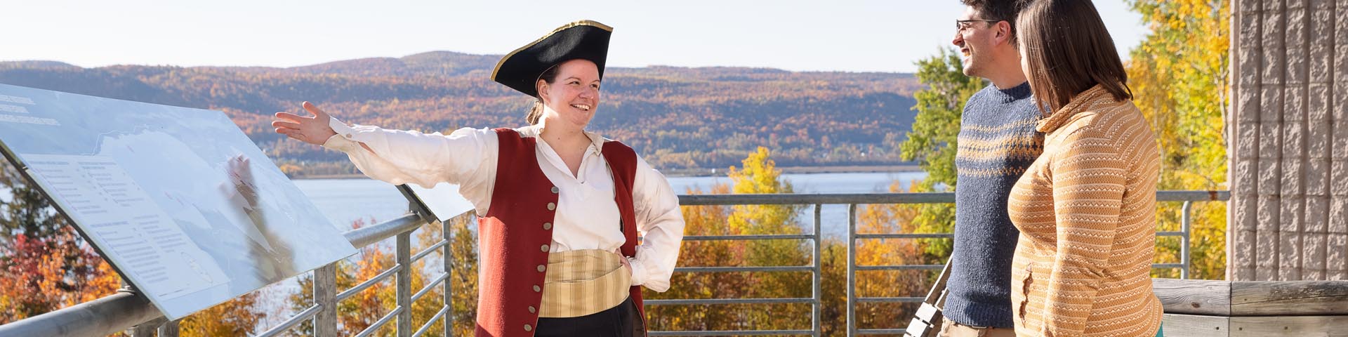 A historical figure talks with visitors against the backdrop of the Restigouche River.