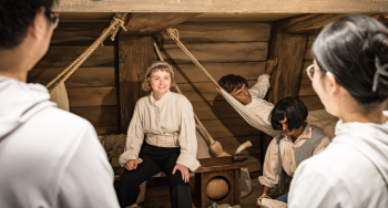 Deux visiteurs de dos regardent une employée costumée en matelot dans le décor d’une cale de navire d’un musée.