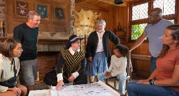 Sitting around a table at Port-Royal National Historic Site a Parks Canada costumed interpreter discusses a map with visitors.